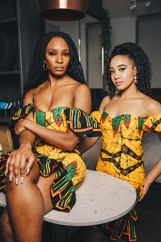 Two ladies one say on a table and other standing. Both are wearing a yellow multi print off the shoulder midi dress with black frills at the bottom. The dresses are made from cotton African print ankara material 
