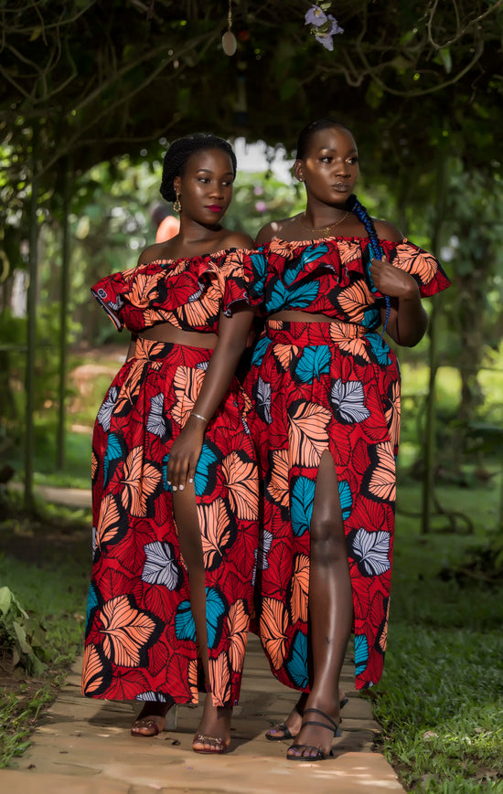 Two ladies standing wearing floral red, blue and orange two piece sets made of high waisted long thigh split skirt and a frill elasticated crop top. The two piece sets are made from cotton African print ankara material 